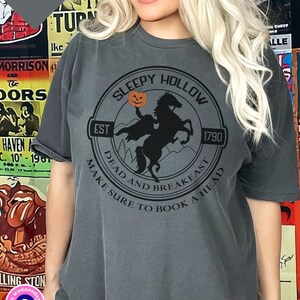 May include: A dark gray t-shirt with a black and white graphic of a headless horseman riding a horse. The graphic is in a circle with the text "Sleepy Hollow" at the top, "Est 1790" at the bottom, and "Dead and Breakfast Make Sure to Book a Head" around the outside.