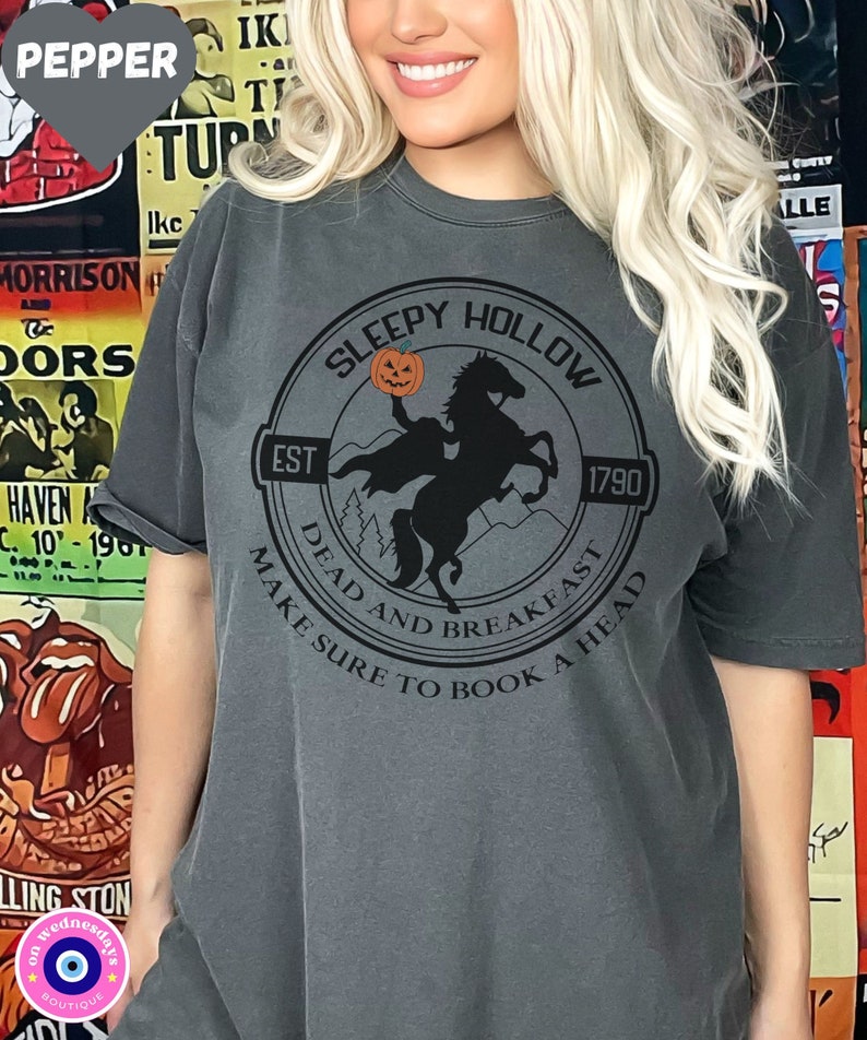 May include: A dark gray t-shirt with a black and white graphic of a headless horseman riding a horse. The graphic is in a circle with the text "Sleepy Hollow" at the top, "Est 1790" at the bottom, and "Dead and Breakfast Make Sure to Book a Head" around the outside.