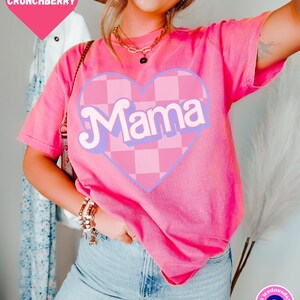 Puede incluir: Camiseta rosa con un coraz&oacute;n a cuadros y la palabra "Mama" en una fuente retro.