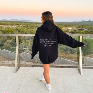 K&ouml;nnte beinhalten: Schwarzer Kapuzenpullover mit dem Text "THE TIMING OF YOUR LIFE WILL MAKE SENSE ONE DAY." Der Hoodie hat eine Kapuze und lange &Auml;rmel. Die Person tr&auml;gt wei&szlig;e Turnschuhe.