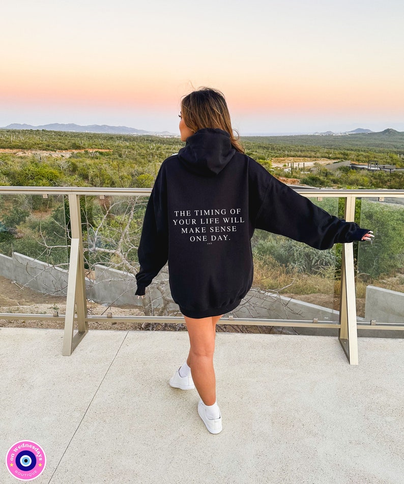 K&ouml;nnte beinhalten: Schwarzer Kapuzenpullover mit dem Text "THE TIMING OF YOUR LIFE WILL MAKE SENSE ONE DAY." Der Hoodie hat eine Kapuze und lange &Auml;rmel. Die Person tr&auml;gt wei&szlig;e Turnschuhe.