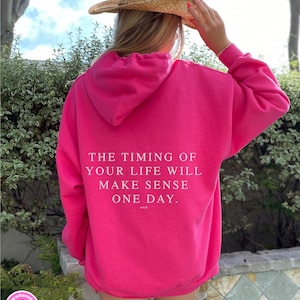 K&ouml;nnte beinhalten: Ein leuchtend pinkfarbener Kapuzenpullover mit dem wei&szlig;en Text "THE TIMING OF YOUR LIFE WILL MAKE SENSE ONE DAY." Der Kapuzenpullover hat eine Kapuze und wird von einer Person getragen, die einen Strohhut tr&auml;gt. Das Boutique-Logo ist unten links sichtbar.