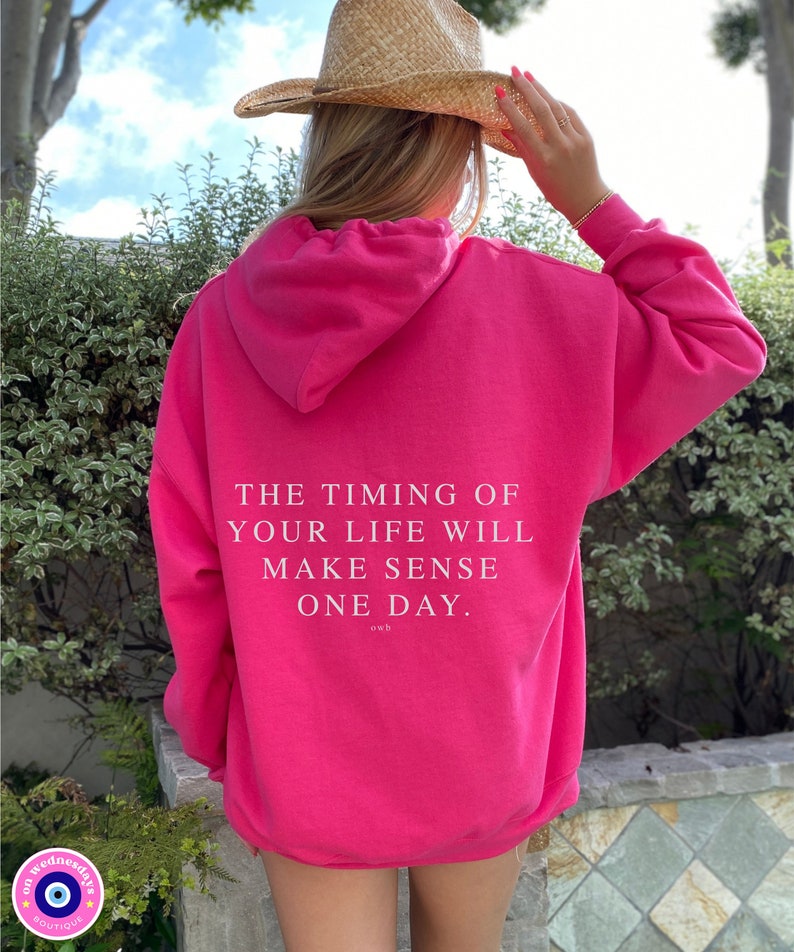 K&ouml;nnte beinhalten: Ein leuchtend pinkfarbener Kapuzenpullover mit dem wei&szlig;en Text "THE TIMING OF YOUR LIFE WILL MAKE SENSE ONE DAY." Der Kapuzenpullover hat eine Kapuze und wird von einer Person getragen, die einen Strohhut tr&auml;gt. Das Boutique-Logo ist unten links sichtbar.