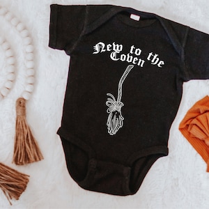 May include: Black baby bodysuit with the text "New to the Coven" and a broom graphic in white. A rust-colored bow headband and a white pumpkin are also in the image. The bodysuit is short-sleeved.