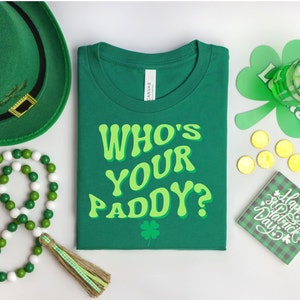 May include: Green t-shirt with the text "Who's Your Paddy?" in a retro font. The shirt is surrounded by St. Patrick's Day themed items, including a green hat, shamrocks, and gold coins.