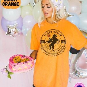May include: Orange t-shirt with a graphic of a black silhouette of a rider on a horse with a pumpkin in its mouth. The graphic is in a circle with the text "Sleepy Hollow" at the top, "Est 1790" at the bottom left, and "Dead and Breakfast Make Sure to Book a Head" around the bottom.