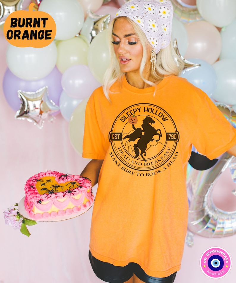 May include: Orange t-shirt with a graphic of a black silhouette of a rider on a horse with a pumpkin in its mouth. The graphic is in a circle with the text "Sleepy Hollow" at the top, "Est 1790" at the bottom left, and "Dead and Breakfast Make Sure to Book a Head" around the bottom.