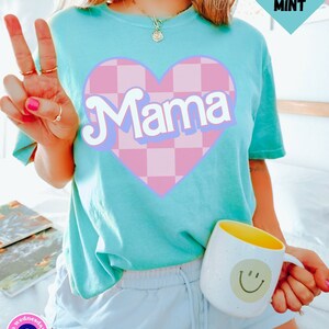 Puede incluir: Una camiseta turquesa con un coraz&oacute;n a cuadros rosa y blanco que dice "Mama" en una fuente retro. La camiseta la lleva puesta una persona con un pantal&oacute;n corto azul claro.