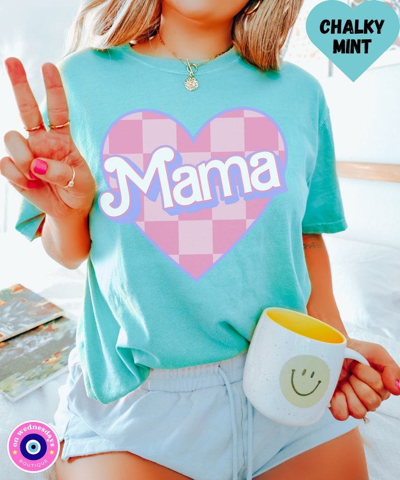 Puede incluir: Una camiseta turquesa con un coraz&oacute;n a cuadros rosa y blanco que dice "Mama" en una fuente retro. La camiseta la lleva puesta una persona con un pantal&oacute;n corto azul claro.