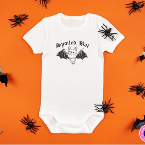 May include: White baby onesie with a black bat graphic and the text "Spoiled Bat" in a gothic font. The onesie is surrounded by black plastic spiders and bats on an orange background.