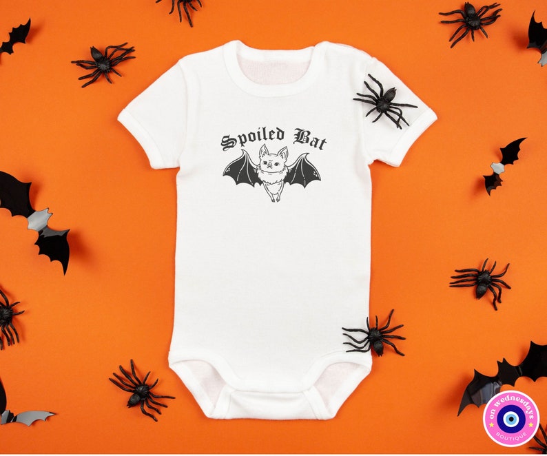 May include: White baby onesie with a black bat graphic and the text "Spoiled Bat" in a gothic font. The onesie is surrounded by black plastic spiders and bats on an orange background.