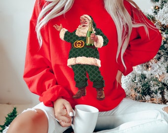 Puede incluir: Suéter rojo de Navidad con un Papá Noel verde y dorado sosteniendo una botella de champán. Papá Noel lleva un traje estampado verde y dorado con una hebilla de cinturón dorada.