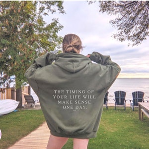 K&ouml;nnte beinhalten: Olivgr&uuml;ner Kapuzenpullover mit dem Text "THE TIMING OF YOUR LIFE WILL MAKE SENSE ONE DAY." Das Bild zeigt den Pullover im Freien mit einem See und B&auml;umen im Hintergrund.