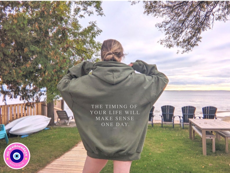 K&ouml;nnte beinhalten: Olivgr&uuml;ner Kapuzenpullover mit dem Text "THE TIMING OF YOUR LIFE WILL MAKE SENSE ONE DAY." Das Bild zeigt den Pullover im Freien mit einem See und B&auml;umen im Hintergrund.