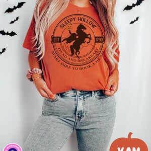 May include: Orange t-shirt with a black and white graphic of a headless horseman riding a horse. The graphic is in a circle with the text "Sleepy Hollow" at the top, "Est 1790" at the bottom right, and "Dead and Breakfast Make Sure to Book a Head" around the bottom.