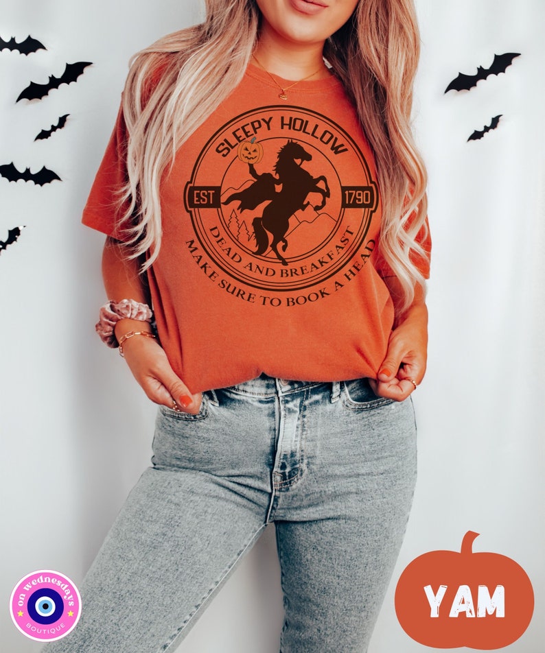 May include: Orange t-shirt with a black and white graphic of a headless horseman riding a horse. The graphic is in a circle with the text "Sleepy Hollow" at the top, "Est 1790" at the bottom right, and "Dead and Breakfast Make Sure to Book a Head" around the bottom.