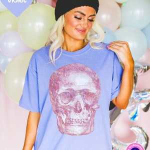 Op de afbeelding: Een lichtblauw T-shirt met een grote, regenboogkleurige glitter-schedelprint. Het shirt heeft een ronde hals en korte mouwen. De tekst "on wednesdays" en een blauw boze oog symbool zijn zichtbaar in de rechter benedenhoek van de afbeelding.