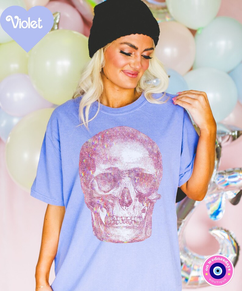 Op de afbeelding: Een lichtblauw T-shirt met een grote, regenboogkleurige glitter-schedelprint. Het shirt heeft een ronde hals en korte mouwen. De tekst "on wednesdays" en een blauw boze oog symbool zijn zichtbaar in de rechter benedenhoek van de afbeelding.