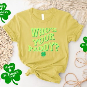 May include: Yellow t-shirt with the text "WHO'S YOUR PADDY?" in green letters. The shirt has a shamrock design and is surrounded by shamrock-shaped decorations with the text "Happy St. Patrick's Day!"