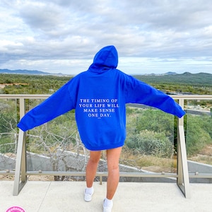 K&ouml;nnte beinhalten: Ein k&ouml;nigsblauer Kapuzenpullover mit dem Text "THE TIMING OF YOUR LIFE WILL MAKE SENSE ONE DAY" auf dem R&uuml;cken. Der Hoodie wird mit wei&szlig;en Turnschuhen getragen. Der Hintergrund zeigt eine malerische Aussicht auf Berge und das Meer.