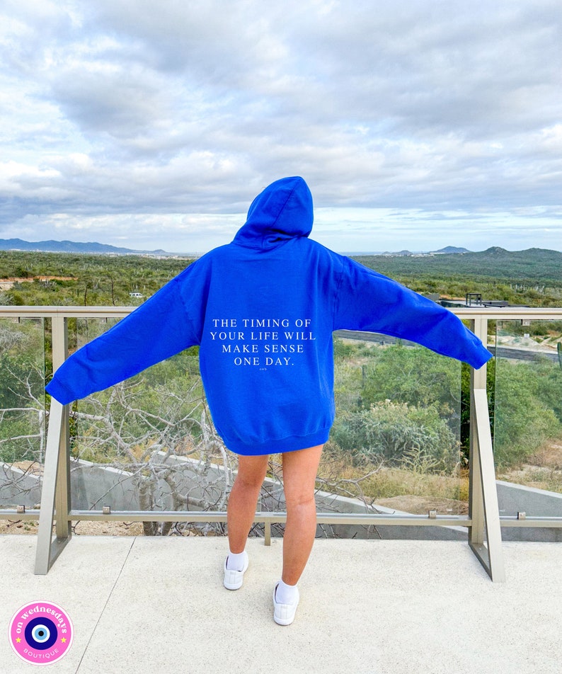 K&ouml;nnte beinhalten: Ein k&ouml;nigsblauer Kapuzenpullover mit dem Text "THE TIMING OF YOUR LIFE WILL MAKE SENSE ONE DAY" auf dem R&uuml;cken. Der Hoodie wird mit wei&szlig;en Turnschuhen getragen. Der Hintergrund zeigt eine malerische Aussicht auf Berge und das Meer.