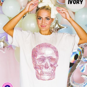 Op de afbeelding: Wit T-shirt met een roze glitter schedel print. De tekst "IVORY" staat in een spraakballon boven de schedel. Het T-shirt heeft een klein logo linksonder dat "on wednesdays boutique" zegt met een boze oog symbool.