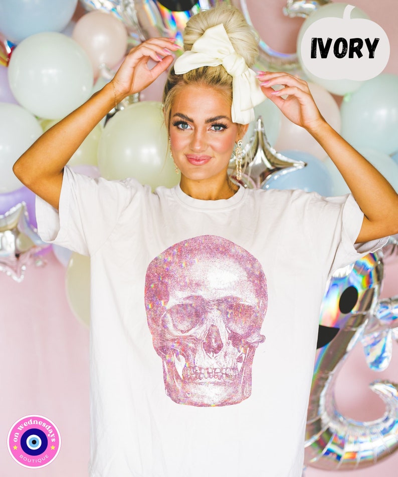 Op de afbeelding: Wit T-shirt met een roze glitter schedel print. De tekst "IVORY" staat in een spraakballon boven de schedel. Het T-shirt heeft een klein logo linksonder dat "on wednesdays boutique" zegt met een boze oog symbool.