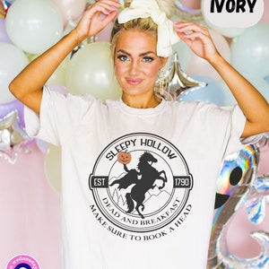 May include: White t-shirt with a black and white graphic of a headless horseman riding a horse with a pumpkin in its hand. The graphic is in a circle with the text "Sleepy Hollow" at the top, "Est 1790" at the bottom left, and "Dead and Breakfast Make Sure to Book a Head" around the bottom.