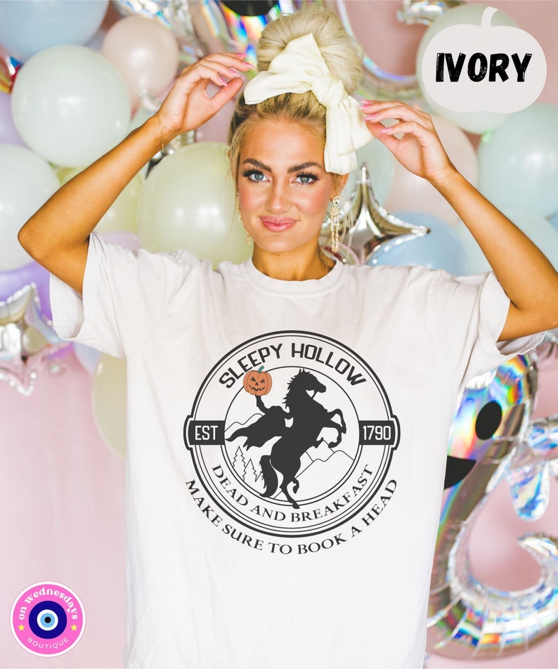May include: White t-shirt with a black and white graphic of a headless horseman riding a horse with a pumpkin in its hand. The graphic is in a circle with the text "Sleepy Hollow" at the top, "Est 1790" at the bottom left, and "Dead and Breakfast Make Sure to Book a Head" around the bottom.
