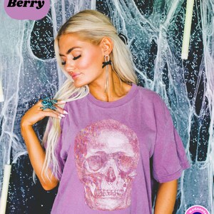 Op de afbeelding: Een paars T-shirt met een glinsterende witte schedelprint. Het shirt heeft een ronde hals en korte mouwen. De tekst "on wednesdays" en een blauw boze oog symbool zijn zichtbaar aan de onderkant van het shirt.