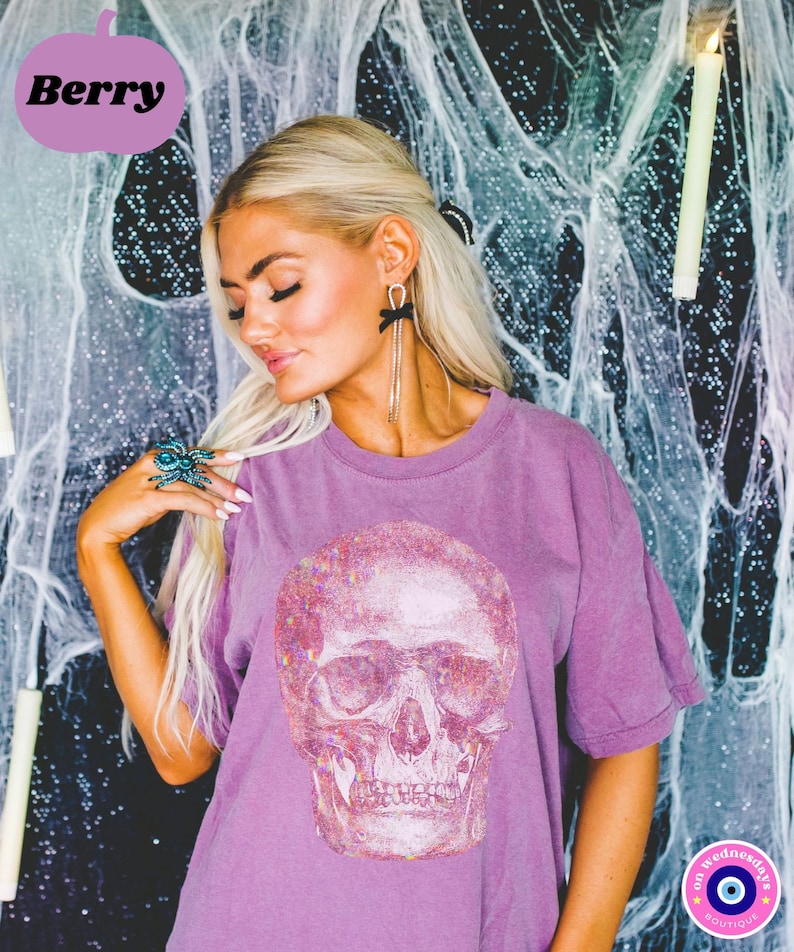 Op de afbeelding: Een paars T-shirt met een glinsterende witte schedelprint. Het shirt heeft een ronde hals en korte mouwen. De tekst "on wednesdays" en een blauw boze oog symbool zijn zichtbaar aan de onderkant van het shirt.