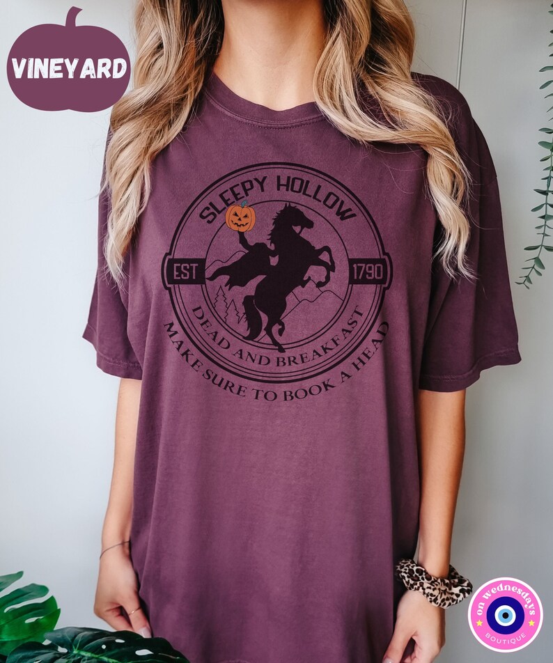 May include: A purple t-shirt with a black graphic of a headless horseman riding a horse. The graphic includes the text "Sleepy Hollow", "Est 1790", "Dead and Breakfast", and "Make Sure to Book a Head".
