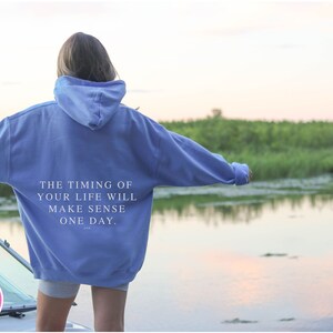 K&ouml;nnte beinhalten: Hellblauer Kapuzenpullover mit dem Text "THE TIMING OF YOUR LIFE WILL MAKE SENSE ONE DAY" auf dem R&uuml;cken. Der Hoodie wird im Freien in der N&auml;he eines Gew&auml;ssers getragen.