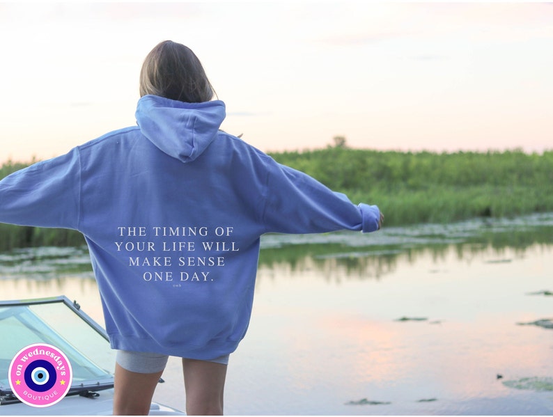 K&ouml;nnte beinhalten: Hellblauer Kapuzenpullover mit dem Text "THE TIMING OF YOUR LIFE WILL MAKE SENSE ONE DAY" auf dem R&uuml;cken. Der Hoodie wird im Freien in der N&auml;he eines Gew&auml;ssers getragen.