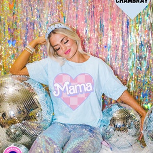 Puede incluir: Una camiseta azul claro con un coraz&oacute;n a cuadros rosa y blanco que dice "Mama" en una fuente retro. La modelo lleva la camiseta con pantalones de lentejuelas y una diadema.