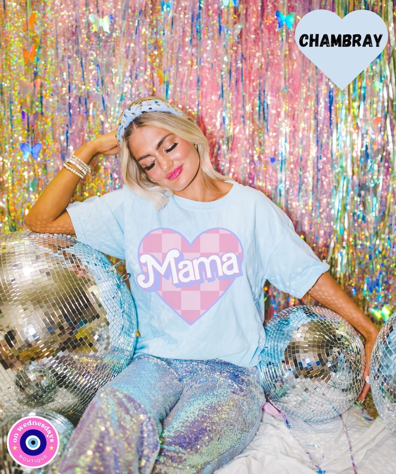 Puede incluir: Una camiseta azul claro con un coraz&oacute;n a cuadros rosa y blanco que dice "Mama" en una fuente retro. La modelo lleva la camiseta con pantalones de lentejuelas y una diadema.