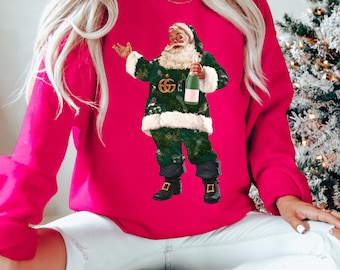 Puede incluir: Una sudadera rosa con un gráfico de Santa Claus verde y blanco que lleva un diseño inspirado en Gucci y sostiene una botella de champán. Santa Claus está de pie sobre un fondo rojo.