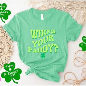May include: A green t-shirt with the text "WHO'S YOUR PADDY?" in yellow letters. The shirt has a shamrock design on the front. There are three shamrock-shaped stickers with the text "Happy St. Patrick's Day!" around the shirt.