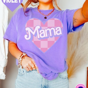 Puede incluir: Una camiseta morada con un coraz&oacute;n a cuadros rosa y blanco que dice "Mama" en una fuente retro.