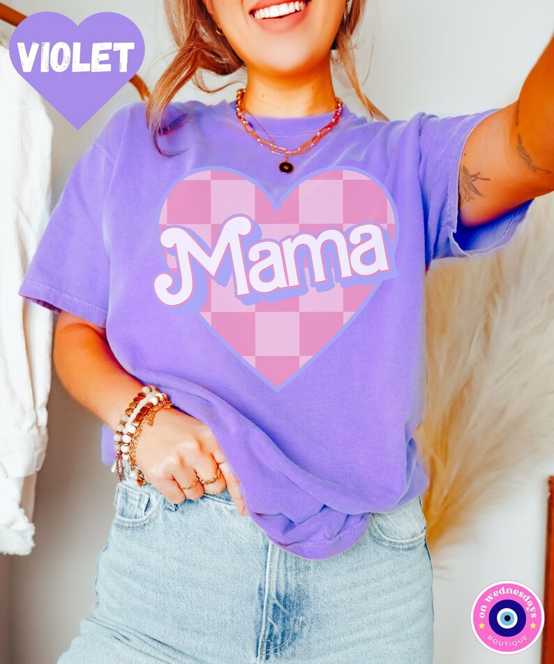 Puede incluir: Una camiseta morada con un coraz&oacute;n a cuadros rosa y blanco que dice "Mama" en una fuente retro.