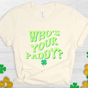 May include: A light beige t-shirt with the text "Who's Your Paddy?" in green letters. The letters have a retro, 70s style. A small green four-leaf clover is below the text.