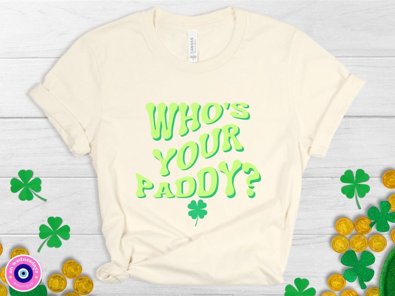 May include: A light beige t-shirt with the text "Who's Your Paddy?" in green letters. The letters have a retro, 70s style. A small green four-leaf clover is below the text.