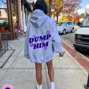Puede incluir: Una sudadera gris con un gráfico morado que dice "DUMP HIM" con flores moradas alrededor del texto. La sudadera tiene capucha y cordón. La persona que lleva la sudadera camina por una acera.