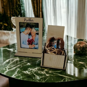 Op de afbeelding: Een houten fotolijst met een foto van een man en een vrouw, de lijst heeft het woord "Mother" erop gegraveerd. Een houten doos met een foto erin staat ook op de tafel.