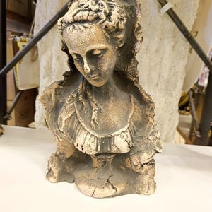 Op de afbeelding: Een verweerde, beige buste van een vrouw met krullend haar en een gedetailleerde, gerimpelde kraag. De sculptuur heeft een klassieke esthetiek, met een serene uitdrukking en een textuur. De buste is een decoratief item.