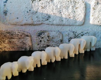 May include: A line of seven white marble elephant figurines of varying sizes, arranged in a row on a black surface. The elephants are facing the same direction, with their trunks raised.