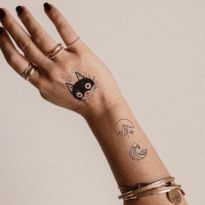 Op de afbeelding: Een arm met tijdelijke tatoeages van een zwarte kat, een kwal en een golf. De persoon draagt gouden armbanden en ringen. De persoon draagt een lichtgekleurde tweedjas.