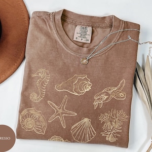 May include: Espresso-colored t-shirt with gold sea life illustrations, including a seahorse, starfish, and seashells. The shirt has a crew neck and a small tag. A silver necklace with a pendant is on the shirt.