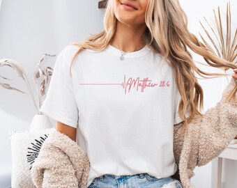 May include: White t-shirt with a pink heartbeat graphic and the text "Matthew 28:6" printed on the front.