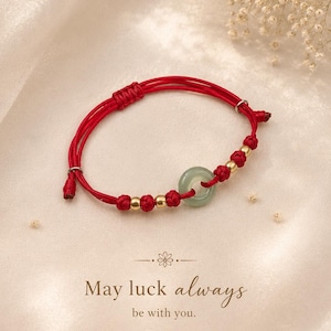 May include: A red cord bracelet featuring a central pale green jade-like ring, accented with small gold-toned metallic beads and knotted details. The text below reads: May luck always be with you. 행운이 언제나 함께하길.